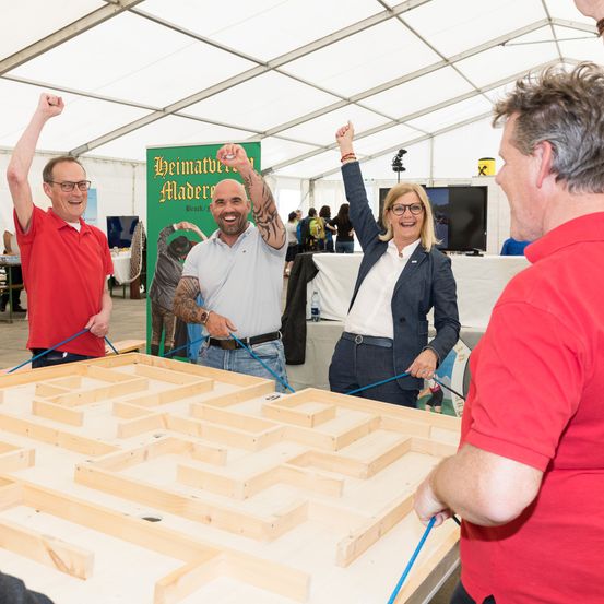 Menschen feiern in einem Zelt. Ein großes Holzlabyrinth liegt auf dem Tisch. Eine Person hält eine blaue Schnur.