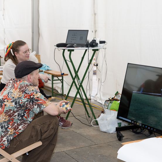 Zwei Personen sind in einem Zelt, eine sitzt und die andere steht, beide benutzen einen Laptop auf einem Tisch mit einem Monitor. Der Mann hält ein Objekt und beide schauen auf den Bildschirm.