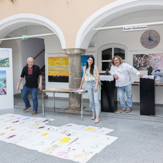 Drei Personen stehen neben einem Tisch mit Kunstwerken in einer Ausstellungshalle. Eine Person trägt ein weißes Hemd, während die anderen zwei lässige Kleidung tragen. Der Boden ist mit zusätzlichen Kunstwerken geschmückt.