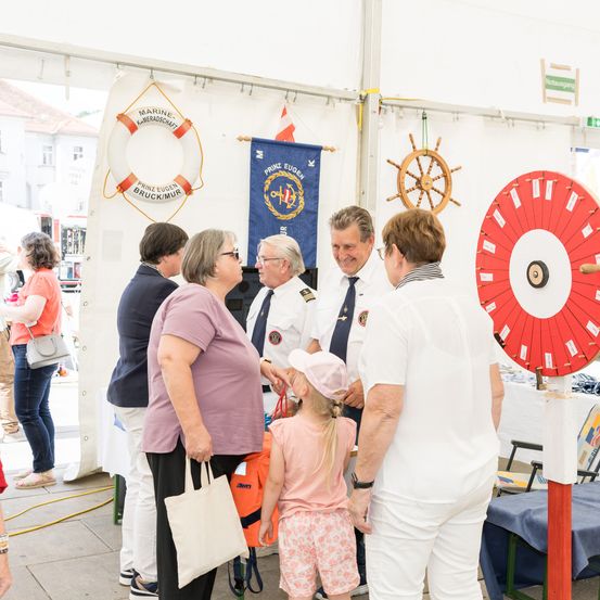 Eine Gruppe von Erwachsenen und einem Kind versammelt sich unter einem Zelt. Sie sind von nautischen Dekorationen umgeben, darunter ein Rettungsring und ein Schiffsrad.