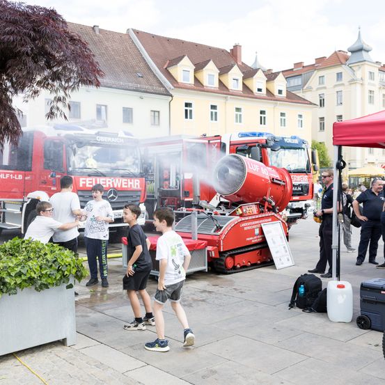 Eine Gruppe von Kindern steht vor einem roten Feuerwehrauto mit einer großen Wassersprüher auf der Straße.