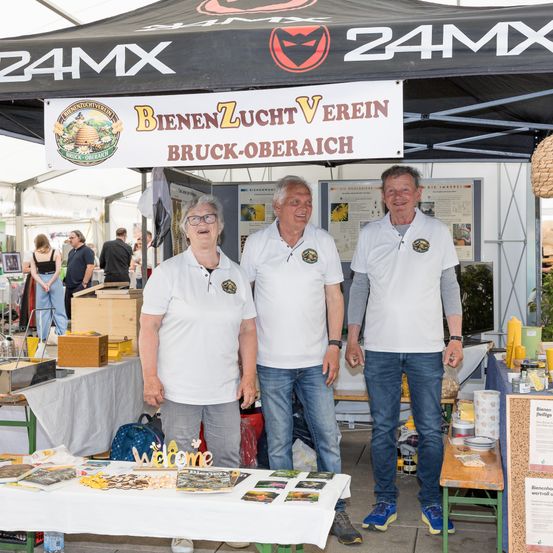 Drei Personen stehen unter einem Zelt mit einem Banner, das 'Bienenzucht Verein Bruck-Oberaich' sagt. Sie tragen weiße Hemden und lächeln. Ein Tisch mit verschiedenen Artikeln steht vor ihnen.