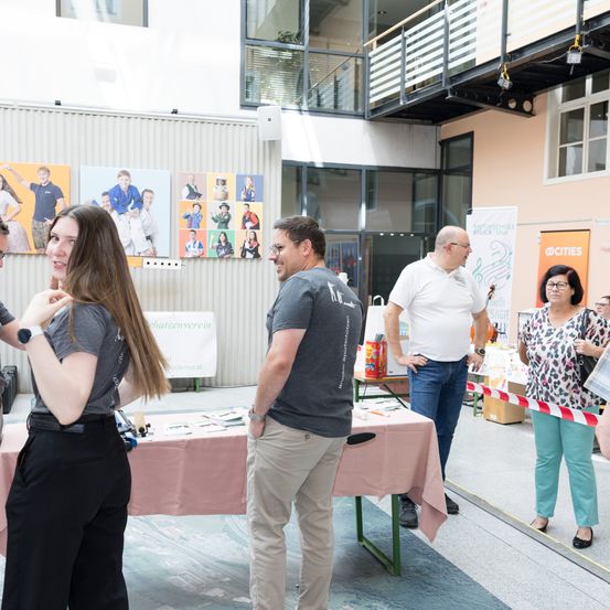 Eine Gruppe von Menschen steht um einen Tisch mit einer rosa Tischdecke herum, möglicherweise bei einer Veranstaltung. Sie schauen sich etwas an, vielleicht eine Ausstellung. Dahinter hängen mehrere Poster an der Wand.