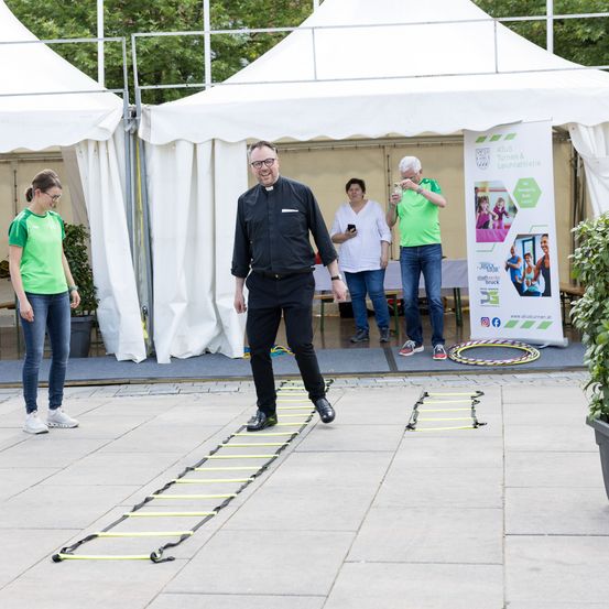 Ein Mann in einem schwarzen Hemd geht auf einer gelben Leiter auf dem Bürgersteig, während zwei Frauen hinter ihm stehen und ein Mann mit einem Hula-Hoop im Hintergrund.