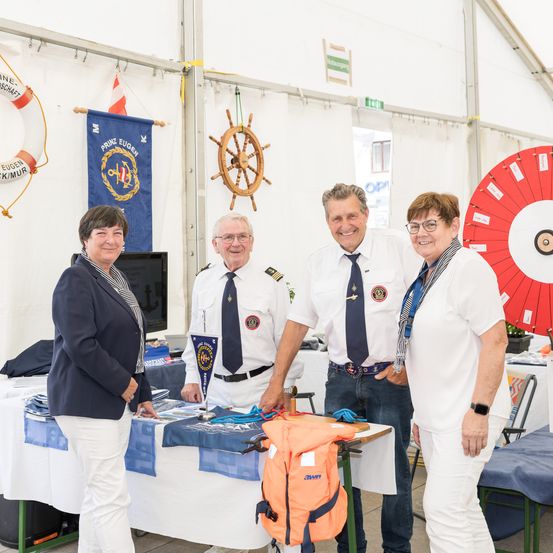 Eine Gruppe von Menschen steht in einem Zelt mit einem Tisch, in Uniformen gekleidet. Ein Mann hält einen Rettungsring und ein Rad. Eine Frau trägt eine Brille, und ein großes Rad befindet sich rechts.