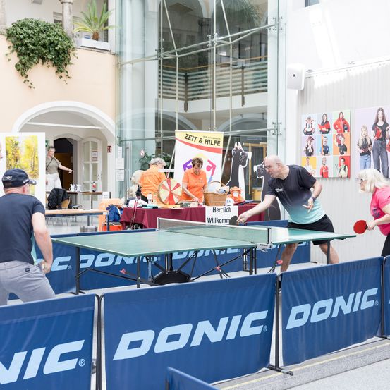 Eine Gruppe von Menschen spielt Tischtennis in einem Außenbereich mit einem blauen Banner und dem Wort Donic. Sie tragen alle Hemden. Es stehen Menschen hinter ihnen. Es gibt einige Tische mit Gegenständen darauf. An der Wand hängt ein Plakat.