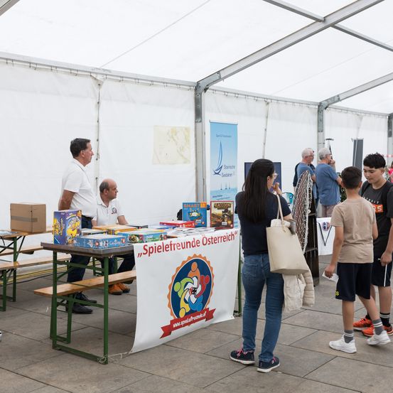Menschen versammeln sich um einen Tisch mit Spielbrettern und Kisten in einem Zelt. Eine Frau hält eine Tragetasche und beobachtet einen Mann mit Rucksack.