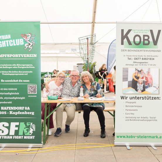 Drei Personen sitzen an einem Tisch mit einem grünen Banner, auf dem 'Tyrolean Fight Club' steht. Sie lächeln und posieren für ein Foto. Ein weiteres Banner dahinter sagt 'Kov' mit den Kontaktdaten der Organisation.
