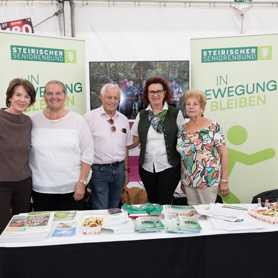 Fünf Personen stehen neben einem Tisch mit Broschüren und Snacks. Zwei Frauen tragen Brillen und Schals, ein Mann hat eine Sonnenbrille an seinem Hemd. Die Banner dahinter lauten 'Steirischer Seniorenbund in Bewegung Bleiben'.