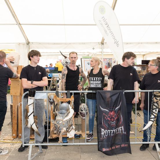 Mehrere Personen stehen hinter einer Barriere bei einer Veranstaltung. Sie tragen schwarze T-Shirts mit Bandlogos. Dahinter zeigt ein Stand verschiedene Gegenstände, darunter Masken und ein Banner mit dem Logo einer Band.
