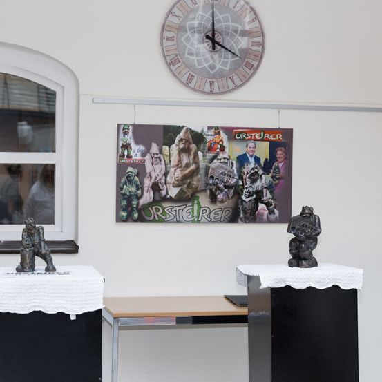 Ein Raum mit zwei Skulpturen auf Ständern, einer Uhr und einem Poster über einem Fenster. Das Fenster spiegelt eine Person in der Ferne wider.