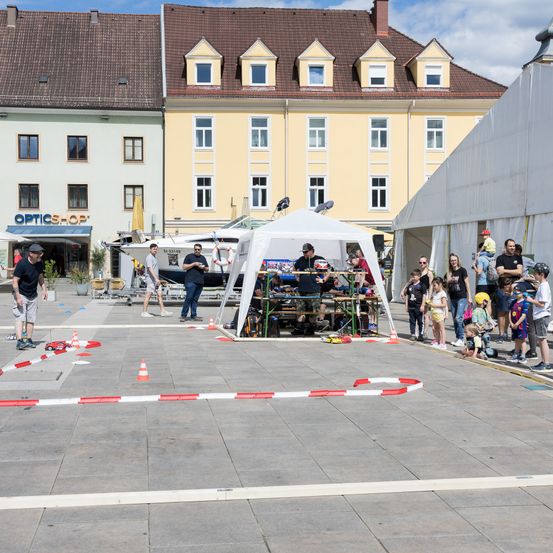 Auf einem offenen Stadtplatz versammeln sich mehrere Personen um eine Rennstrecke mit Zelten und Tischen. Ein Mann fährt ein Auto auf der Strecke, während andere zusehen. Gebäude mit vielen Fenstern umgeben den Bereich.