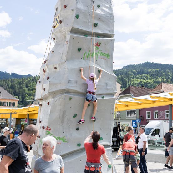 Ein Mädchen in einer pinken Mütze klettert an einer Kletterwand auf einem Stadtplatz. Leute beobachten sie, darunter eine ältere Frau und ein Mann in einem schwarzen T-Shirt. Im Hintergrund befinden sich Gebäude und Berge.