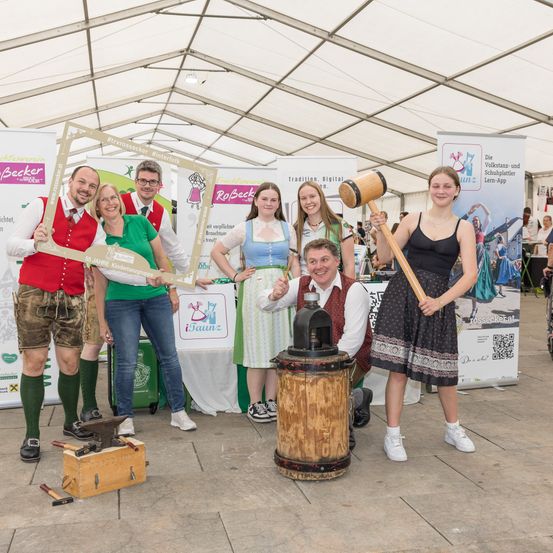 Eine Gruppe von Menschen posiert für ein Foto auf einem Volksfest. Ein Mann und eine Frau halten einen Hammer. Sie stehen neben einem Fass und einem Amboss.