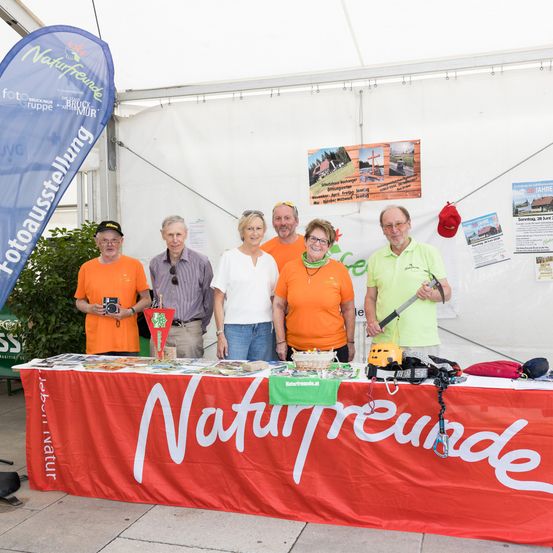 Eine Gruppe von Menschen steht hinter einem Tisch und lächelt für ein Foto. Der Tisch hat einen roten Stoff mit weißen Texten und verschiedene Gegenstände darauf. Sie tragen orangefarbene Hemden. Ein Banner hängt hinter ihnen.