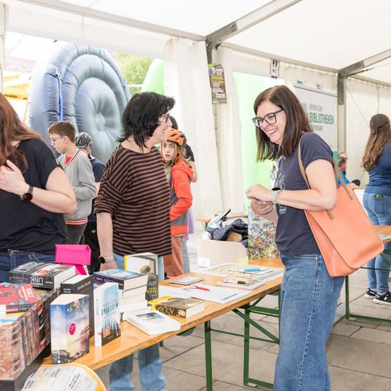 Mehrere Personen stöbern bei einer Freiluft-Buchmesse durch Bücher. Eine Frau mit Brille lächelt. Im Hintergrund sind aufblasbare Rutschen und ein Zelt zu sehen.