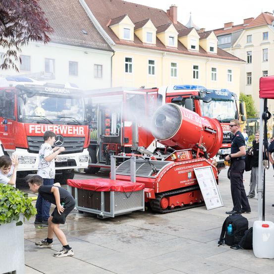 Eine Gruppe von Kindern spielt mit Wasser aus dem Schlauch eines Feuerwehrwagens. Ein Feuerwehrmann steht in der Nähe, und mehrere Feuerwehrwagen sind dahinter geparkt.