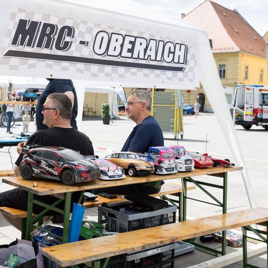 Zwei Männer sitzen unter einem Vordach mit der Aufschrift MRC-Oberaich, mit Modellautos auf einem Tisch. Ein Krankenwagen fährt im Hintergrund.