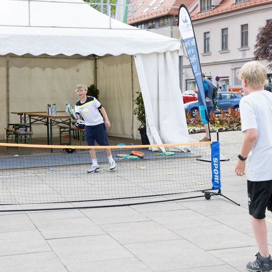 Zwei Jungen spielen Tennis auf einem temporären Platz in einem öffentlichen Bereich. Ein Junge schlägt einen Tennisball, während der andere zuschaut.
