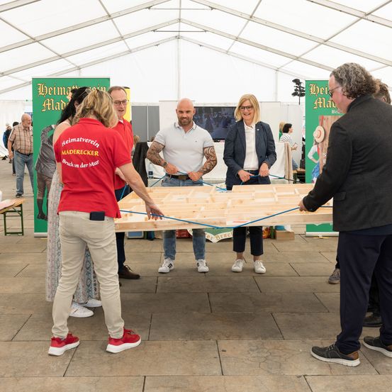 Eine Gruppe von Menschen steht um ein Holzbrett herum und arbeitet möglicherweise an einem Projekt. Eine Frau in Rot hält das Brett, während andere helfen. Die Umgebung scheint ein Ereignis unter einem weißen Zelt zu sein.