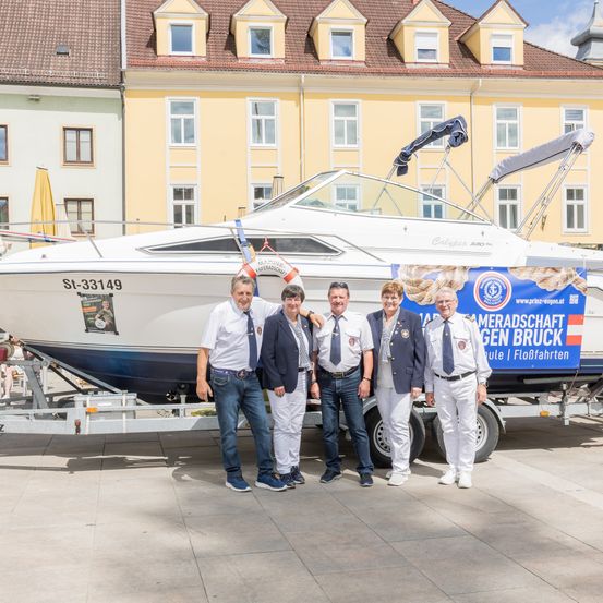 Fünf Personen stehen vor einem weißen Boot auf einem Anhänger. Dahinter befindet sich ein gelbes Gebäude. Eine blaue Fahne ist am Boot befestigt.