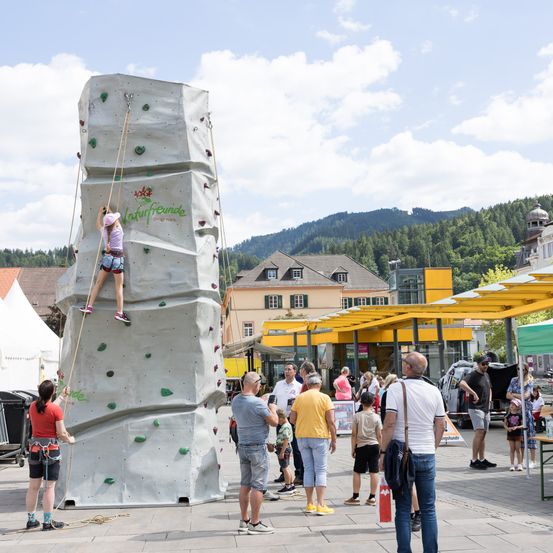Ein Mädchen in einem rosa Shirt klettert an einer Kletterwand auf einem Stadtplatz, umringt von Menschen. Ein Gebäude mit einem gelben Dach ist im Hintergrund, und es gibt Bäume und Berge in der Ferne.