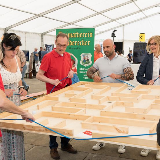 Vier Erwachsene arbeiten zusammen an einem Holzlabyrinth-Spiel in einem Innenraum. Zwei Männer halten Seile, während die Frauen zusehen.
