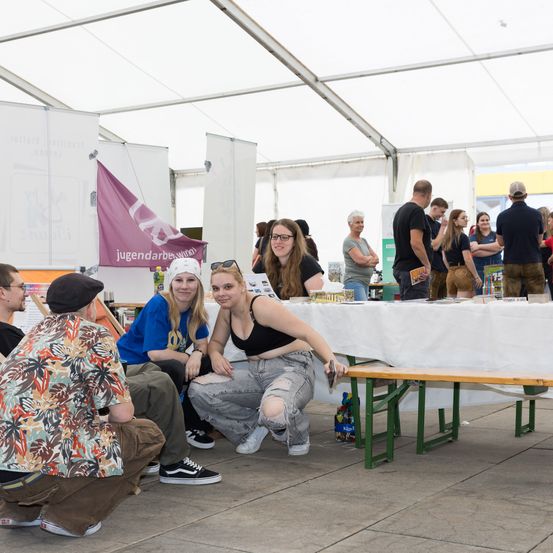 Mehrere Menschen sitzen und stehen in einem Zelt, einige lächeln, unter einem weißen Baldachin. Ein Tisch mit Gegenständen ist mit einem weißen Tuch bedeckt. Eine lila Fahne ist links angebracht.