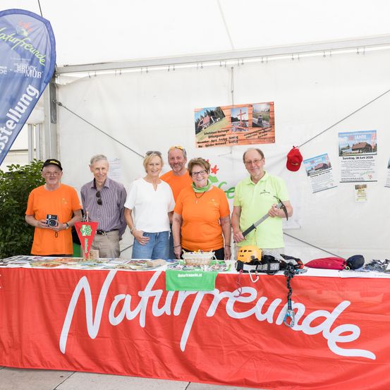 Eine Gruppe von Menschen steht in einem Zelt und posiert für ein Foto. Sie tragen orangefarbene Hemden und halten eine Flagge. Vor ihnen steht ein Tisch mit einem Banner, auf dem Naturfreunde steht.
