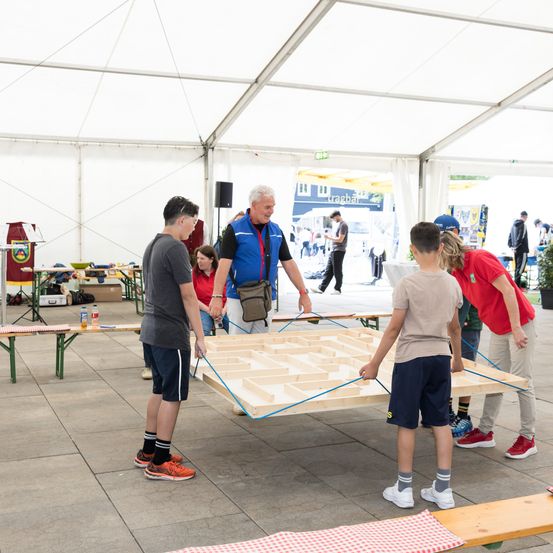 Menschen bewegen eine große Holzplatte in einem Zelt, wobei mehrere Leute beobachten. Ein Mann lächelt und schaut auf die Platte.