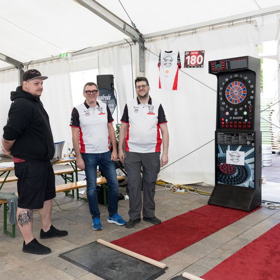 Drei Männer stehen in einem Zelt, einer mit einer Tätowierung am Bein. Sie sind neben einem Dartboard und einem Whiteboard mit der Nummer 180.