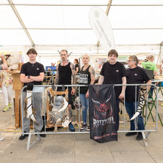 Eine Gruppe von Menschen steht hinter einem Metallzaun. Ein Banner mit einem roten Teufel ist am Zaun befestigt. Ein Mann auf der linken Seite hält einen Becher. Die Leute tragen schwarze T-Shirts.