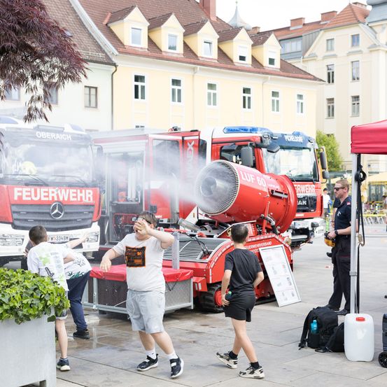 Drei Kinder und ein Feuerwehrmann stehen vor einem roten Feuerwehrauto, das Wasser sprüht. Ein Mann steht unter einem Zelt und ein großes Gebäude ist im Hintergrund.