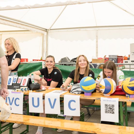 Vier Frauen sitzen unter einem Zelt an einem Tisch mit Volleyballs. Jede Frau hält einen Volleyball und lächelt in die Kamera. Der Tisch zeigt die Buchstaben 'UVC'.