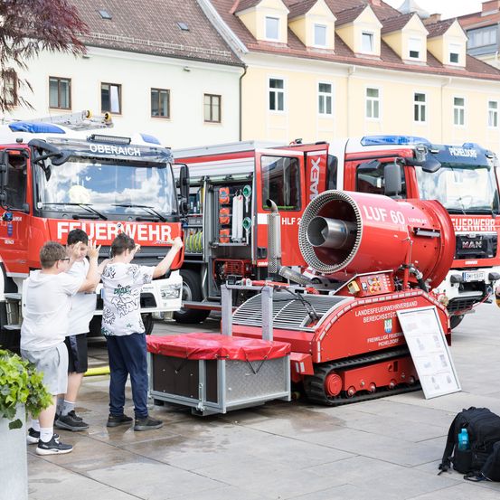 Drei Jungen untersuchen ein rotes Fahrzeug mit der Aufschrift 'LUF 60'. Sie stehen vor mehreren Feuerwehrwagen in einer Stadt.