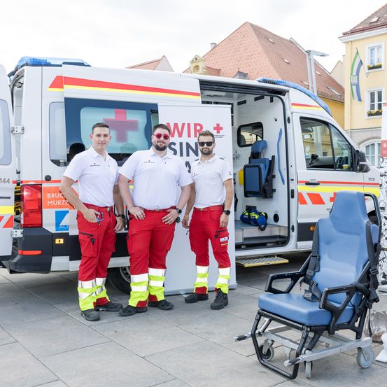 Drei Männer in roten und weißen Uniformen stehen neben einem Krankenwagen mit offener Tür. Ein blauer Stuhl steht neben ihnen.