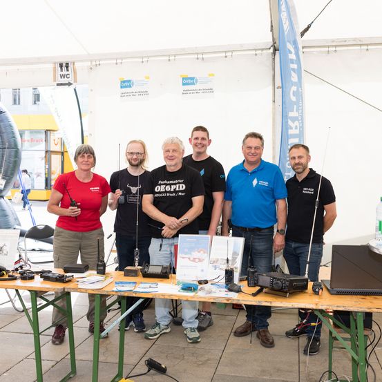 Sechs Personen stehen hinter einem Tisch mit Funkgeräten und lächeln für ein Foto. Ein Banner im Hintergrund steht 'V1K-A'.