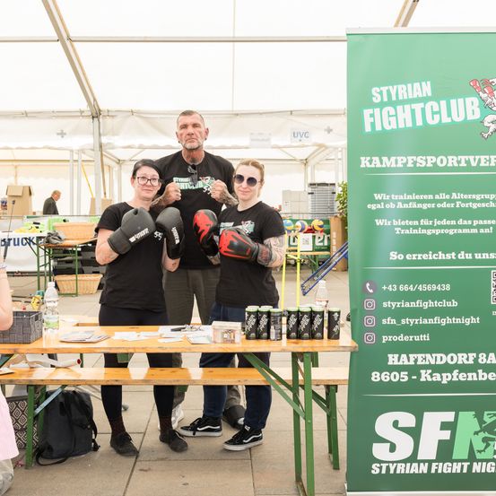 Drei Personen, die Boxhandschuhe und T-Shirts tragen, stehen vor einem Banner, das für den Styrian Fight Club wirbt. Ein Tisch mit Produkten und Flaschen steht vor ihnen.