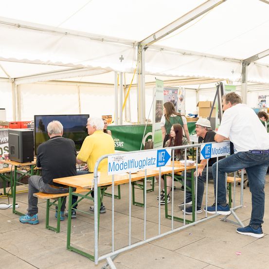 Mehrere Personen sitzen in einem Zelt mit Metallbarrieren. Ein Mann steht und spricht mit einer Frau. Ein Monitor und ein Lautsprecher stehen auf dem Schreibtisch. Banner und Lagerboxen sind dahinter.