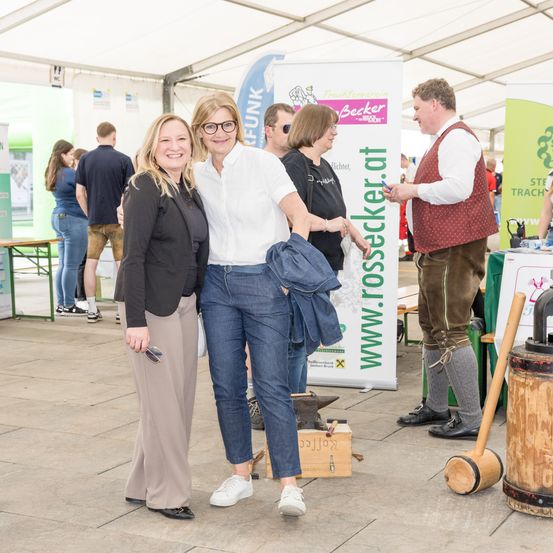Zwei Frauen stehen lächelnd neben einem Mann in traditioneller deutscher Kleidung. Hinter ihnen ist ein Schild mit 'www.rosssecker.at' zu sehen. Andere Menschen sind im Hintergrund, einige in Freizeitkleidung.