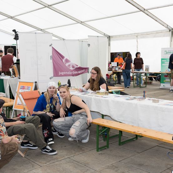 Eine Gruppe von Menschen sitzt unter einem Zelt bei einer Outdoor-Veranstaltung, mit einem langen Tisch vor ihnen, auf dem Bücher und andere Gegenstände ausgestellt sind. Eine Frau posiert für ein Foto.