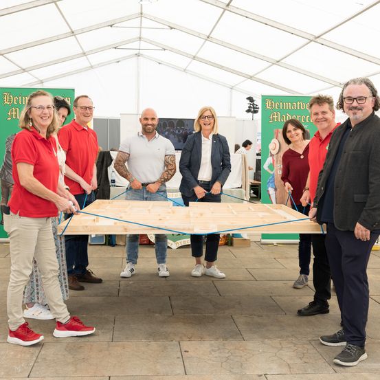 Eine Gruppe von Menschen in roten und weißen Hemden steht um eine große Holzplatte mit blauer Schnur herum. Sie lächeln und posieren für ein Foto. Hinter ihnen sind grüne Banner.