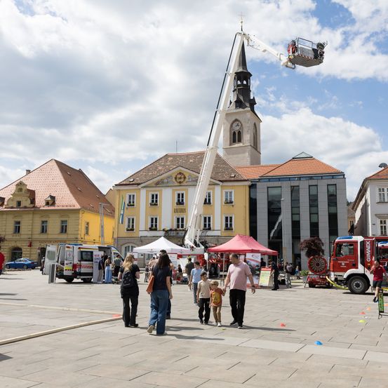 Ein überfüllter Stadtplatz mit Gebäuden, einem Feuerwehrwagen und einer Arbeitsbühne. Mehrere Menschen gehen und einige stehen in der Nähe von Zelten.