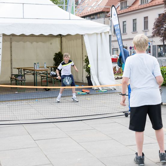 Zwei Jungen spielen Tennis auf einem Platz mit einem Netz vor einem Zelt, ein Junge schlägt den Ball mit einem Schläger.