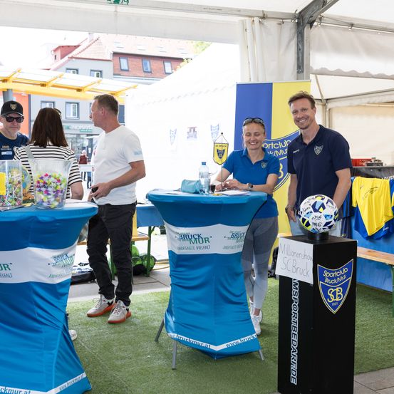 Eine Gruppe von Menschen bei einer Sportveranstaltung, zwei Personen lächeln in die Kamera. Tische mit blauen Abdeckungen und einem Fußball. Ein Banner mit Text hinter den Menschen.
