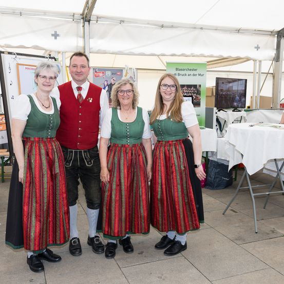 Vier Menschen in traditioneller Kleidung stehen in einem Zelt. Ein Mann in einem roten Vest und drei Frauen in grünen Kleidern mit roten und grünen Streifen. Dahinter ein Tisch mit weißer Tischdecke, ein Monitor und ein Schild.