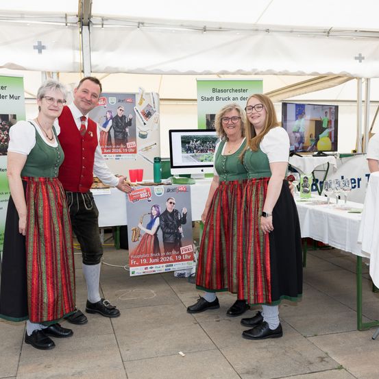 Vier Personen in traditioneller bayerischer Kleidung stehen an einem Zeltstand und halten ein Plakat. Sie lächeln und posieren für ein Foto.