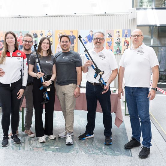 Sechs Menschen stehen zusammen und halten Luftgewehre. Drei Männer und drei Frauen. Sie lächeln und posieren für ein Foto.