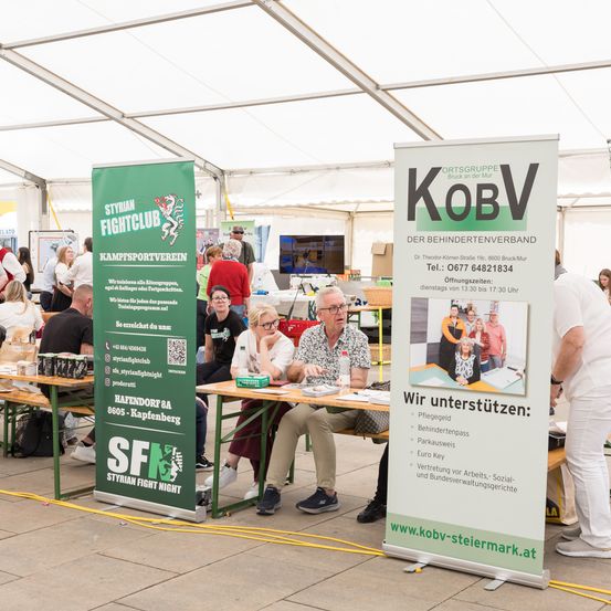 Eine Gruppe von Menschen ist in einem Zelt versammelt. Zwei Männer sitzen mit einer Frau an einem Tisch. Sie scheinen etwas zu besprechen. Banner sind auf der linken und rechten Seite des Bildes aufgestellt. Das Banner auf der rechten Seite hat den Text 'KOBV'.