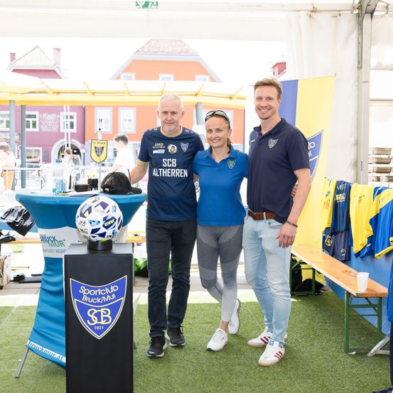Drei Personen stehen neben einem Fußball auf einem Podest und tragen blaue Poloshirts mit einem Logo. Der Mann auf der linken Seite trägt eine Sonnenbrille. Hinter ihnen ist ein Tisch mit einem blauen Banner zu sehen. Im Hintergrund sind Gebäude und eine Bank mit Trikots zu sehen.